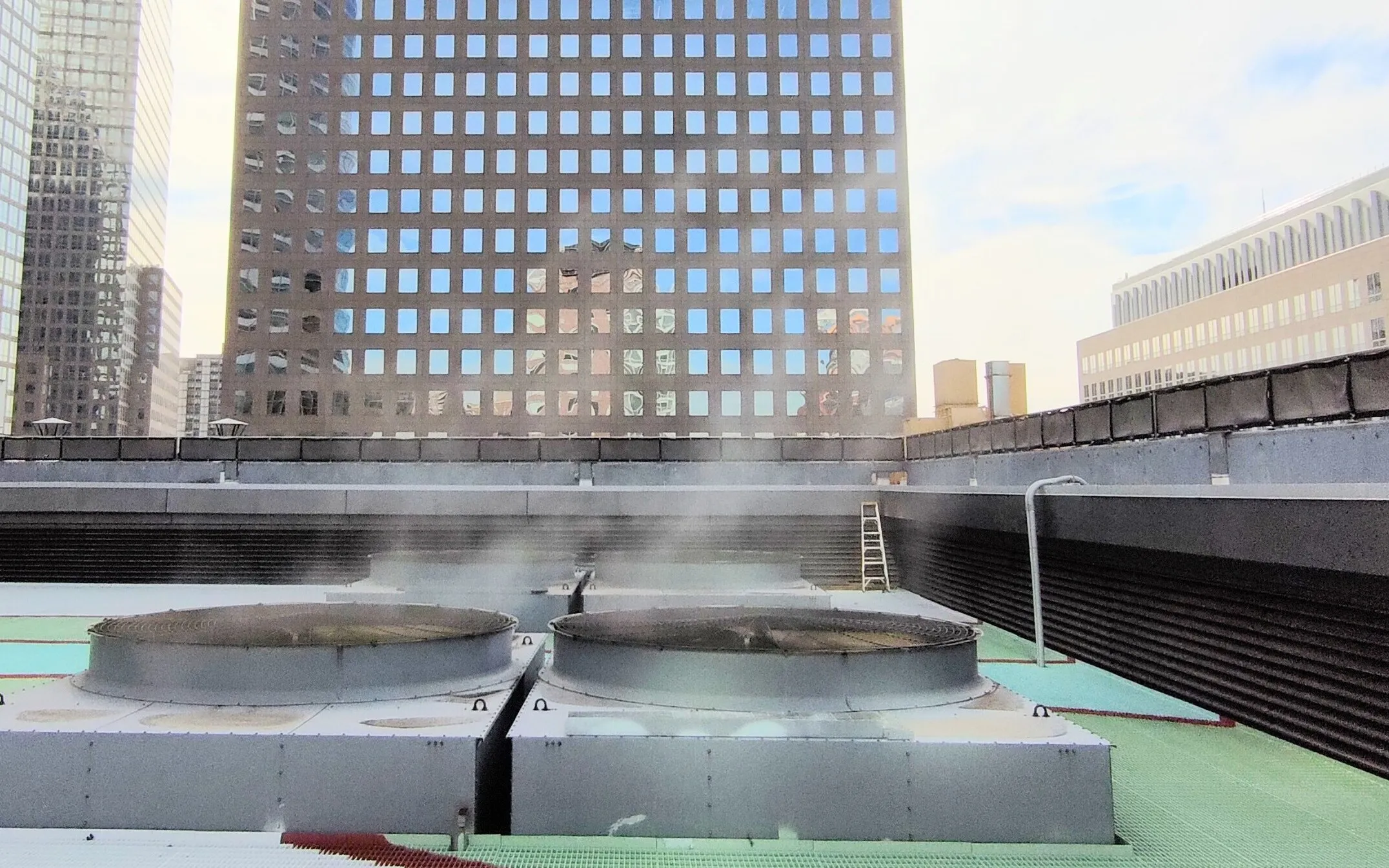 Two rooftop cooling tower fans with a plume of vapour, office towers behind, in Lower Manhattan